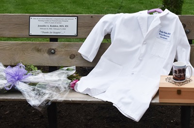 A bench with a memorial plaque for Jennifer Rodden with a white coat and other remembrance items